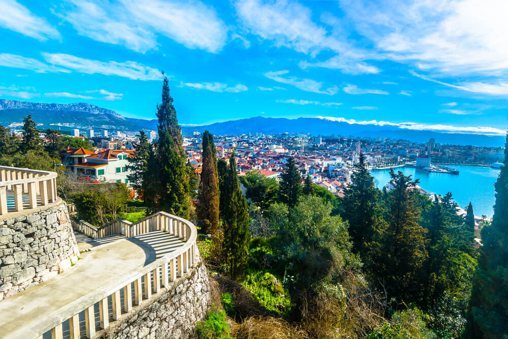 Pohľad na mesto Split a more z chodníka na vrchole meste, kam vedie cesta, ihličnany, jasná obloha a modré more.