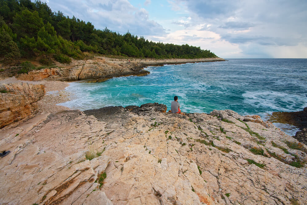 Pohľad na chorvátske pobrežie Kacjak. Na mohutnej skale krémovej farby sedí muž, pozerá sa na búrlivé more tyrkysové more prechádzajúce do modrej farby. Pobrežie je obsypané kamienkami.